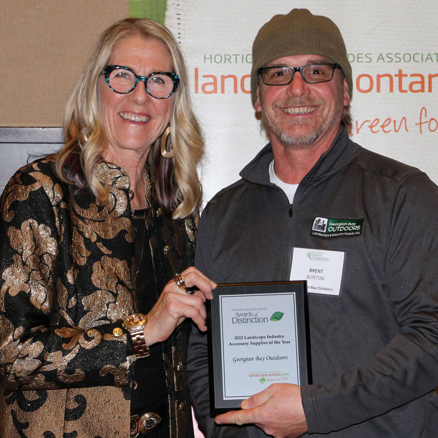Two smiling people holding an award plaque, with a banner in the background related to horticulture or landscaping, at an indoor event.