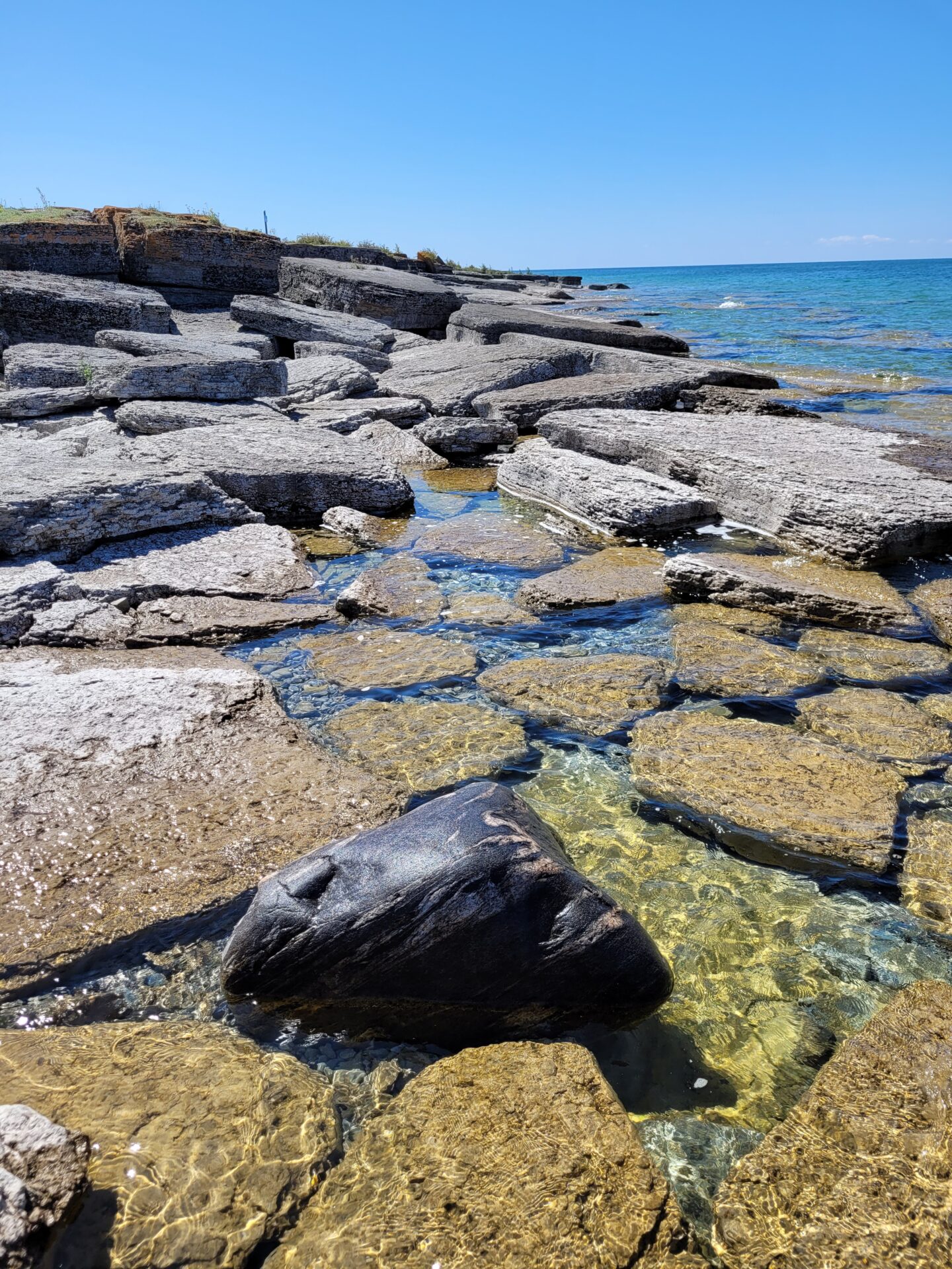 Rocky coastline with clear, shallow water under a bright blue sky. The sunlight highlights textured stones, creating a serene, natural seascape.