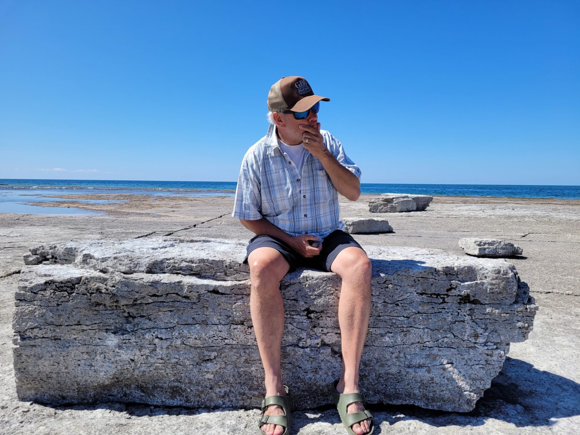 A person in a plaid shirt and cap sits on a rock by the ocean under clear blue skies.