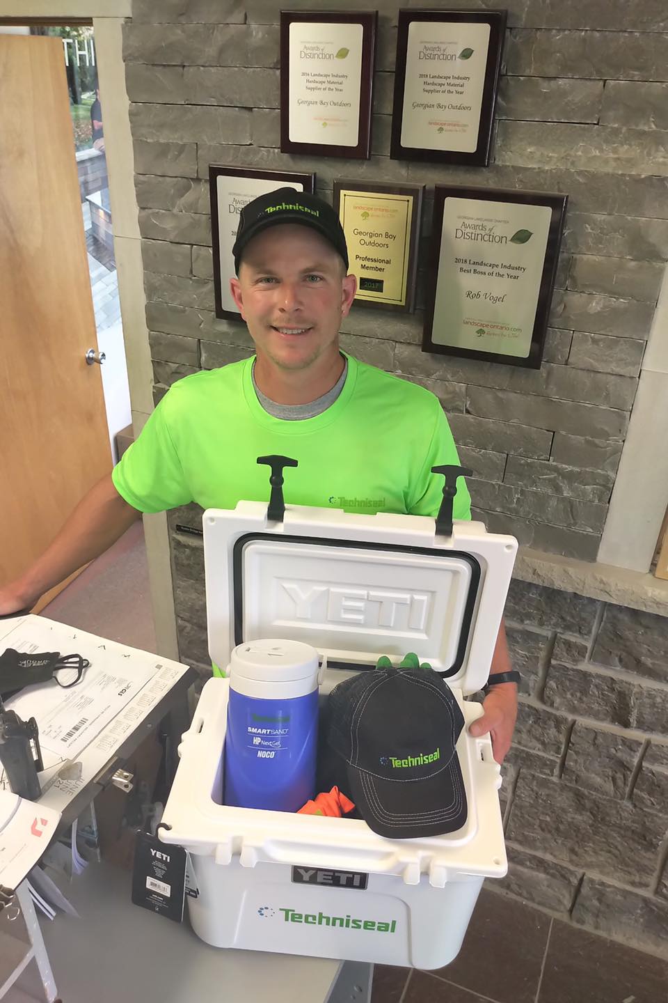 A person in green holds a Yeti cooler with items, standing by award plaques on a stone wall.