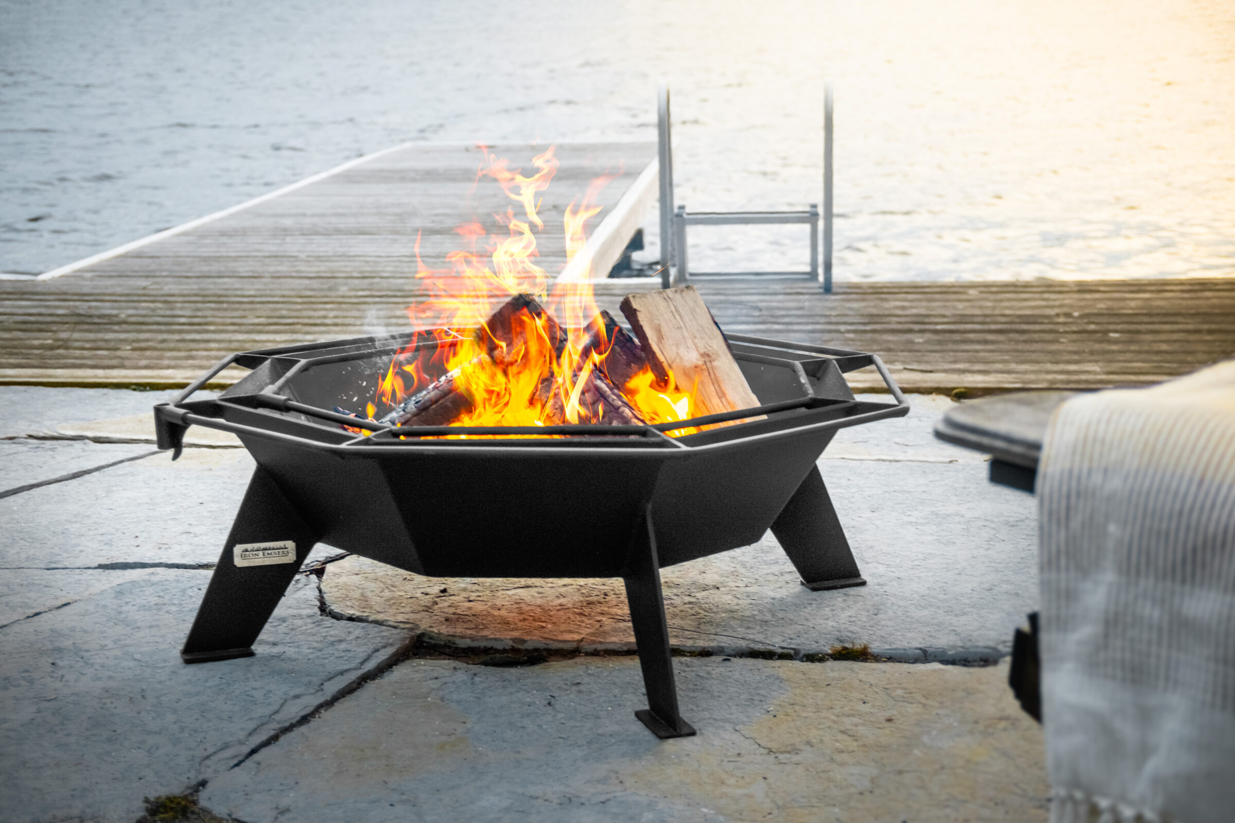 A fire pit burns with bright flames beside a dock leading to a calm lake, creating a cozy outdoor setting.