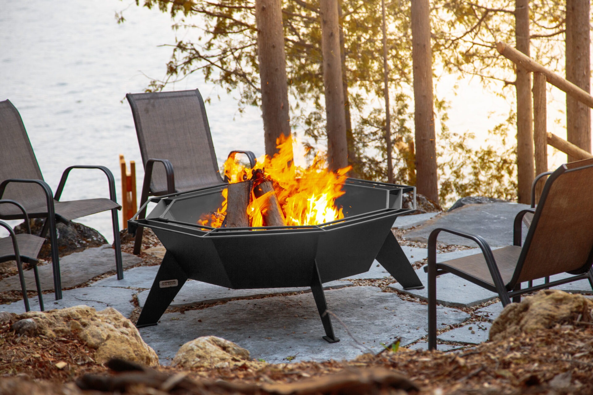 A cozy backyard setup with chairs around a hexagonal fire pit, surrounded by trees and overlooking a calm lakeside.