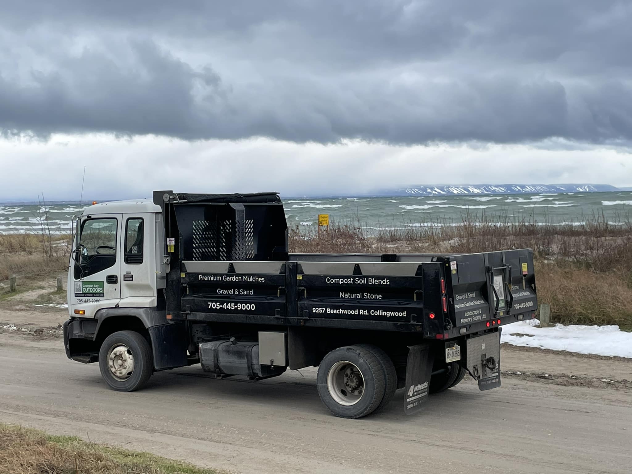 A truck parked on a dirt road near a turbulent lake. Snowy mountains are visible in the distance under a cloudy sky.