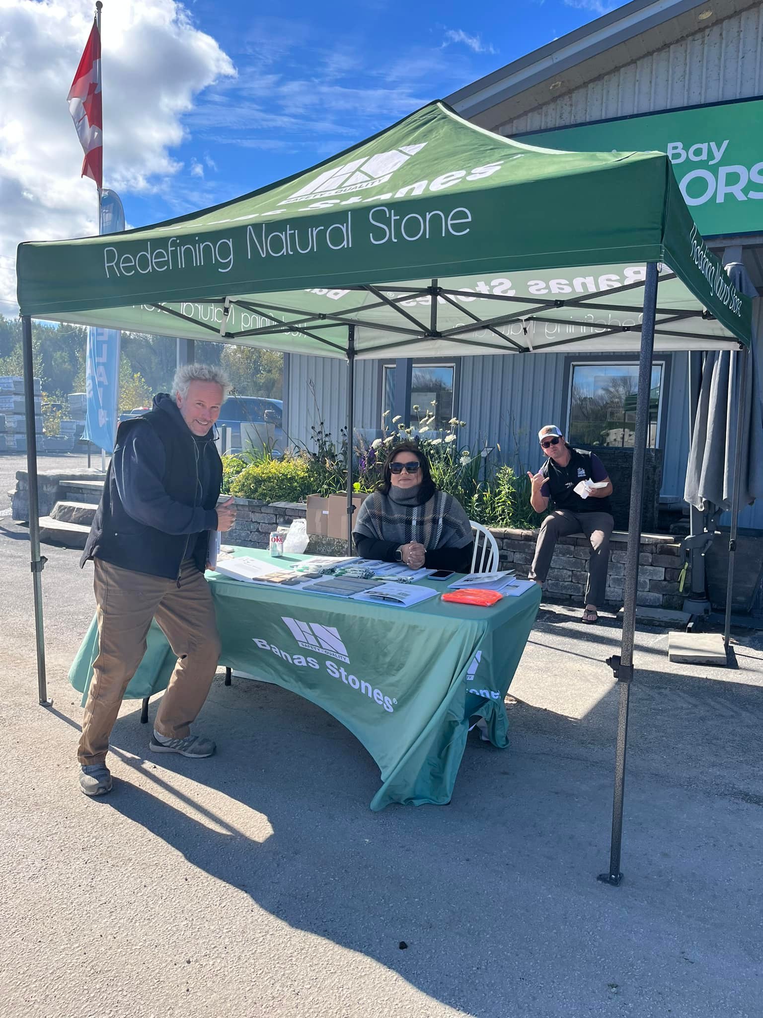 Three people at a Banas Stones booth under a green canopy, with Canadian flag nearby, at an outdoor event under a clear sky.
