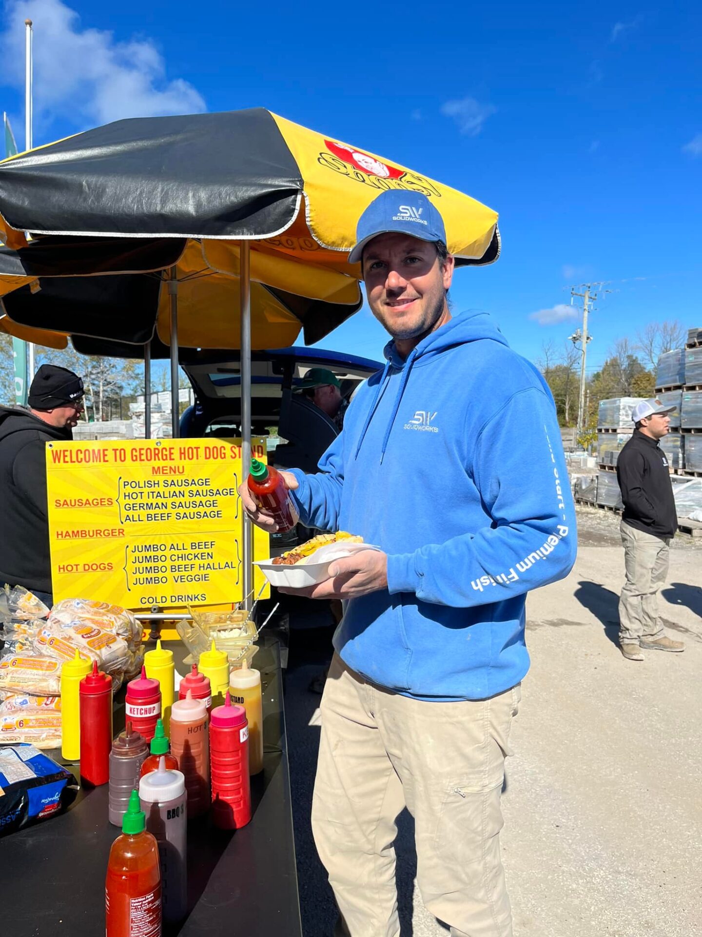 A person in a blue hoodie prepares hot dogs at a stand with various condiments under a yellow umbrella, outdoors.