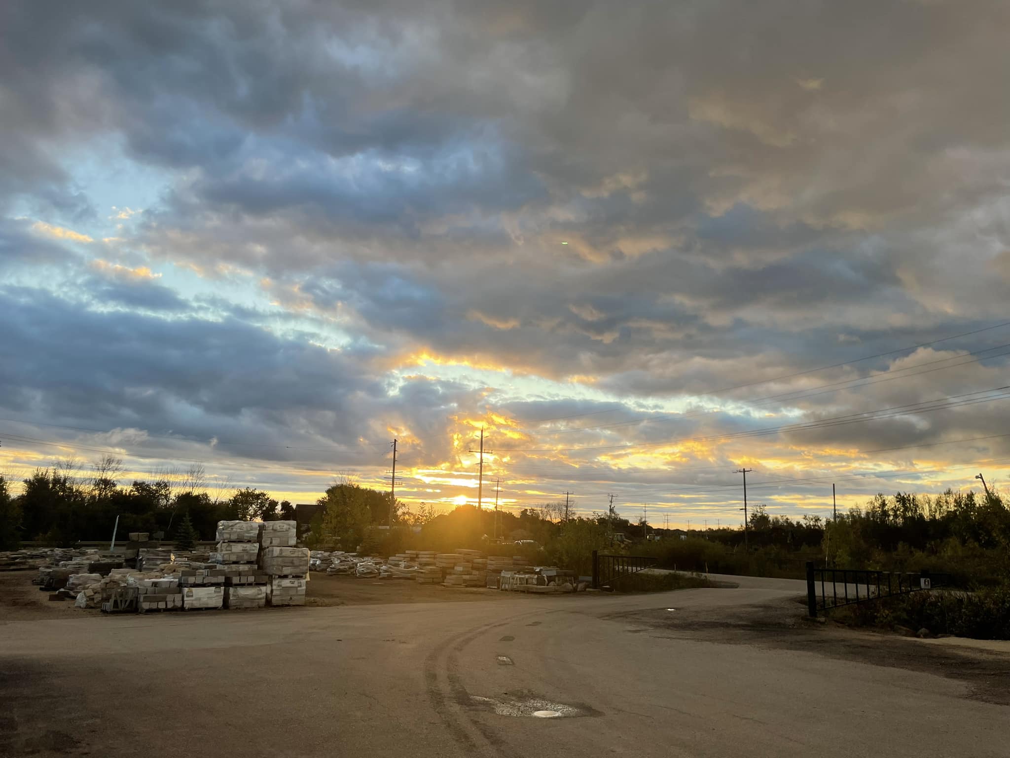 Sunset over a storage yard with stacks of materials, surrounded by trees and power lines, under a dramatic, cloudy sky.
