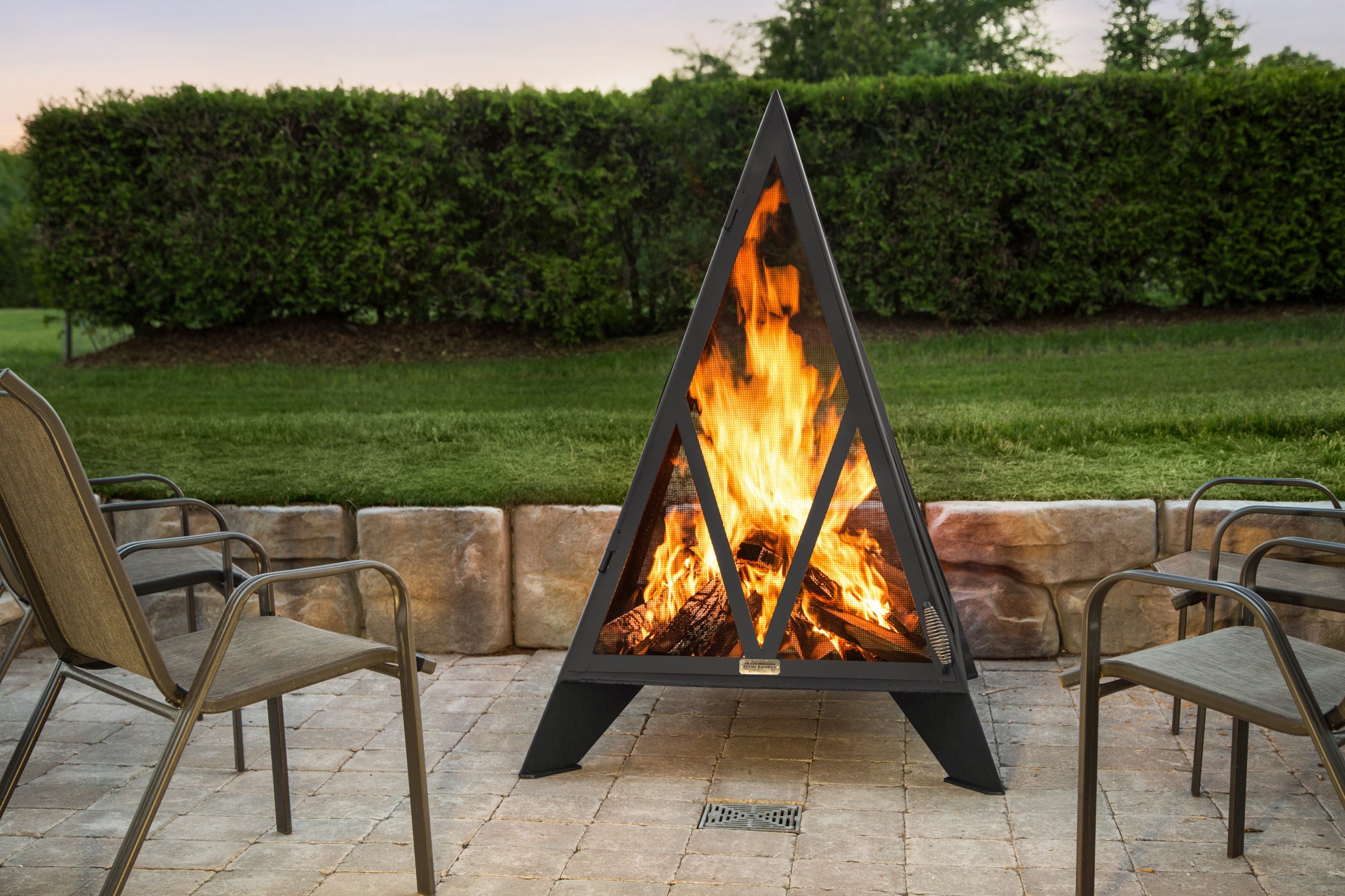 A triangular outdoor fireplace burns brightly on a patio, surrounded by four empty chairs, with a hedge and lawn in the background.