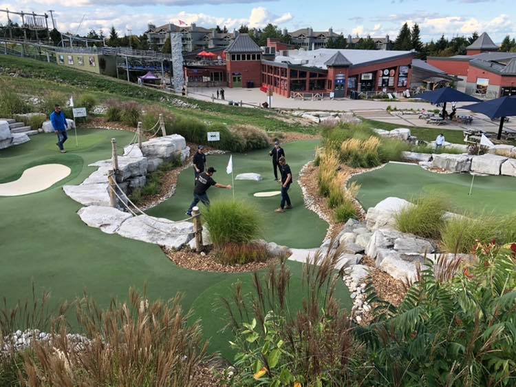 People play mini-golf on a course with artificial grass and rocks. A building complex stands in the background under a cloudy sky.