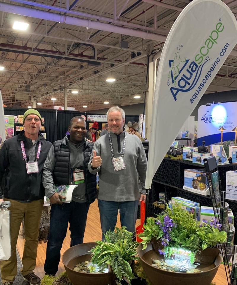 Three people smiling at an indoor trade show booth with Aquascape sign, showcasing water features and plants. Bright lighting and industrial ceiling visible.