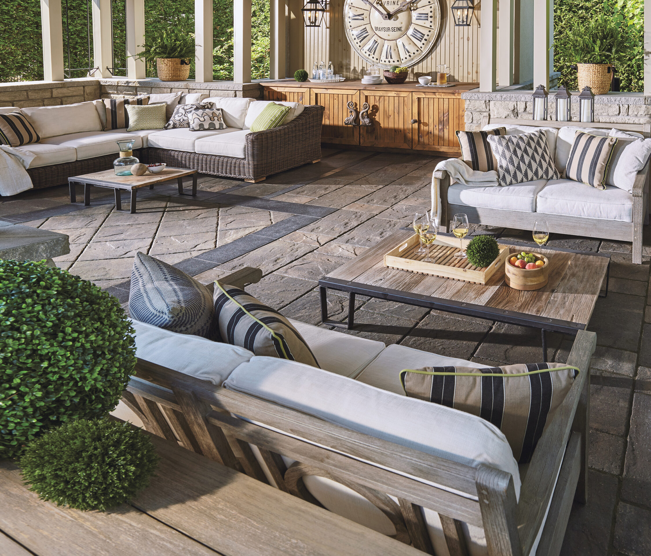 Stylish outdoor patio with cushioned seating, a large decorative clock, wooden tables, and green plants. Wine glasses and snacks on tables.