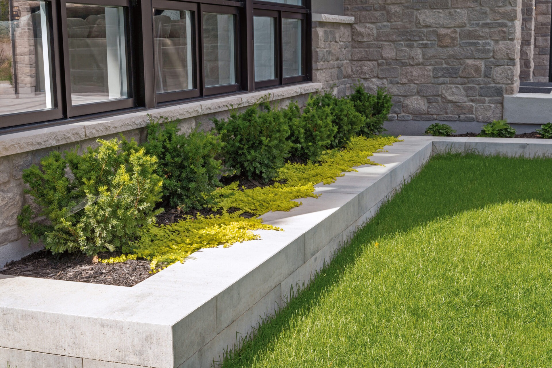 Stone building exterior with modern windows, landscaped with green bushes and yellow ground cover, adjacent to a neatly maintained grassy lawn.