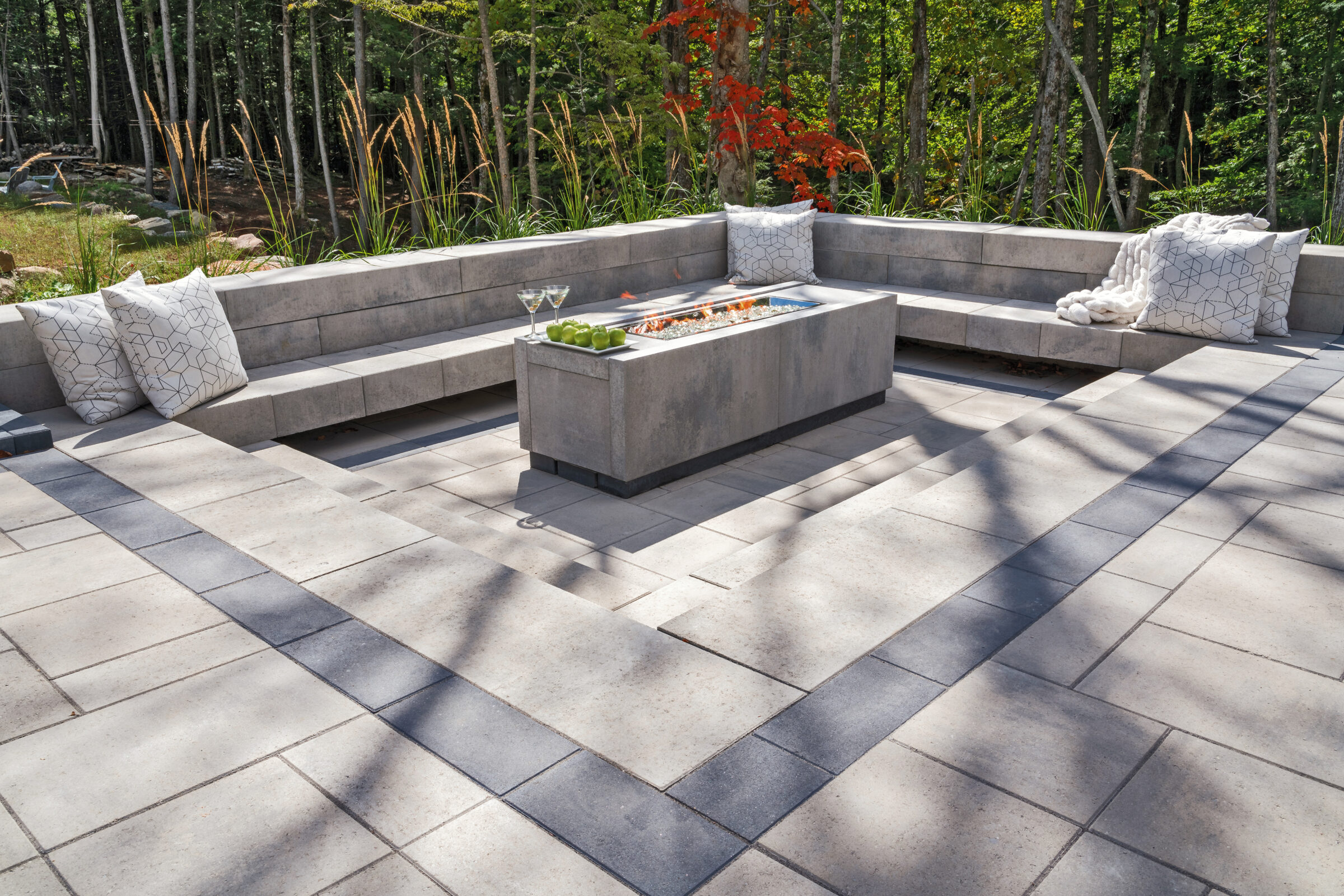 Modern outdoor patio with concrete benches, a fire pit centerpiece, and cushions. Surrounded by trees and foliage for a tranquil setting.
