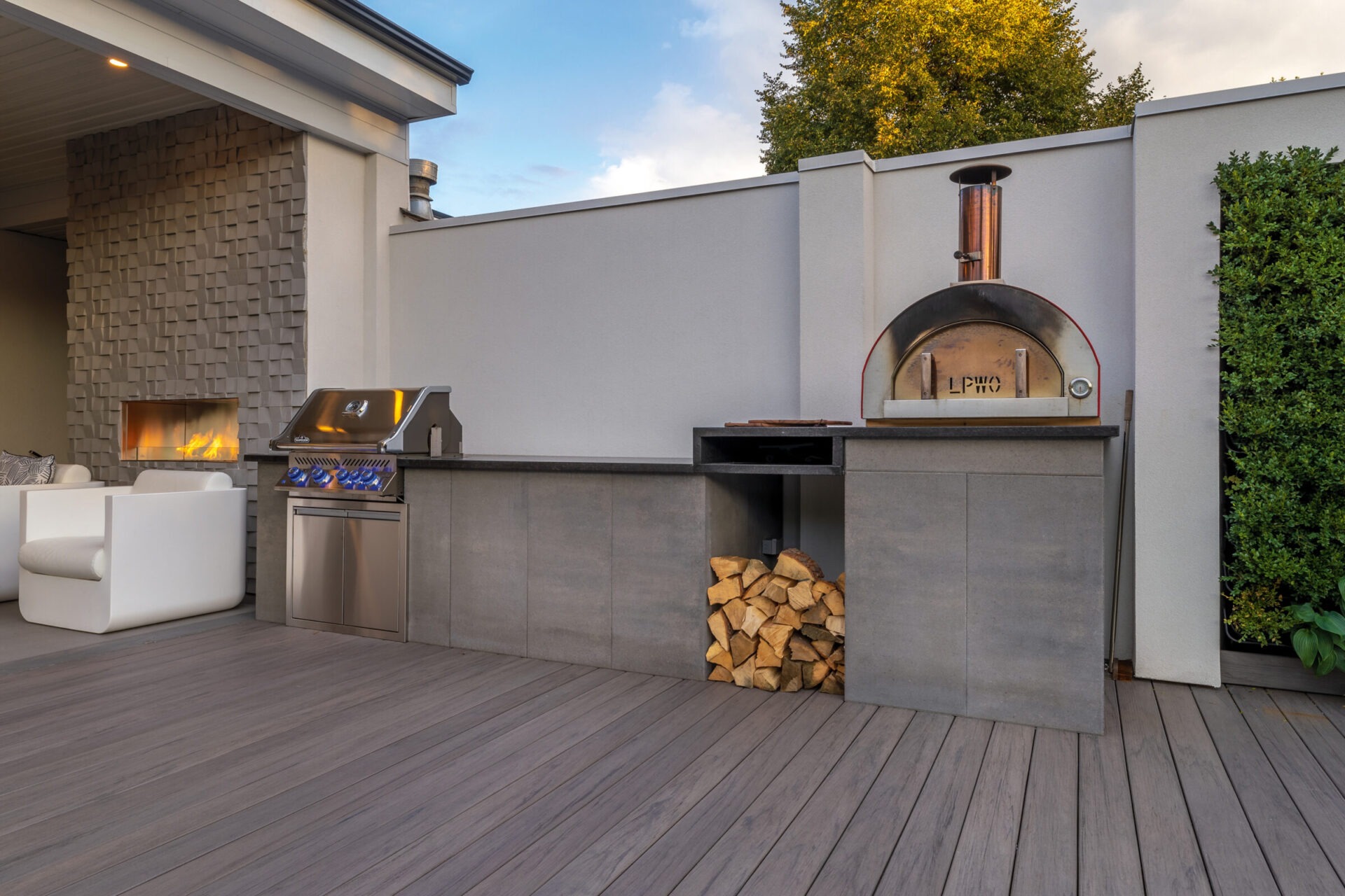 Modern outdoor kitchen with a pizza oven, grill, and stacked firewood. Includes a fireplace and contemporary seating on a wooden deck.
