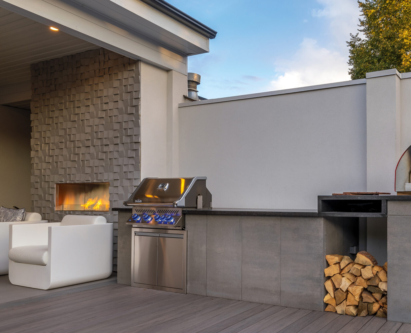 Modern outdoor kitchen with a pizza oven, grill, and stacked firewood. Includes a fireplace and contemporary seating on a wooden deck.
