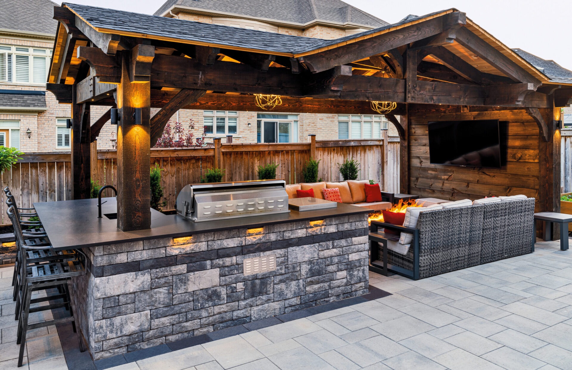 Outdoor patio with stone bar, grill, and seating area. Wooden pergola and firepit create a cozy atmosphere in a residential backyard setting.