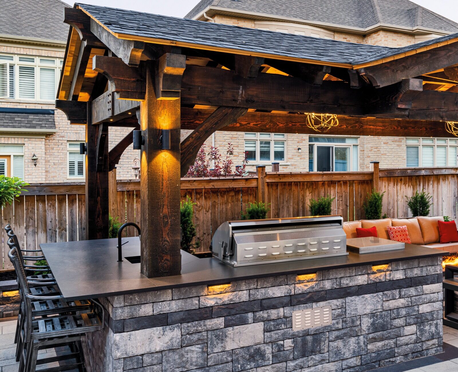 Outdoor patio with wooden pergola, stone bar, grill station, fire pit, and cozy seating area. Modern backyard setup in suburban home.