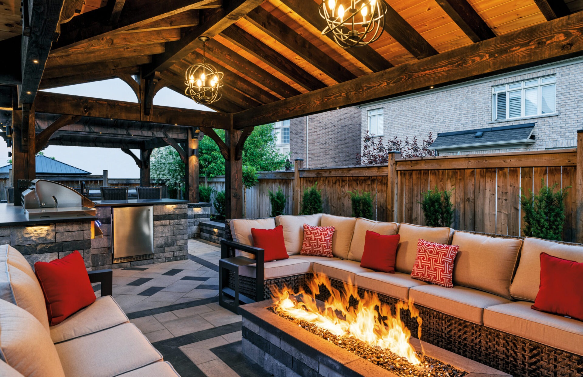 Cozy outdoor patio with wooden pergola, modern grill, and stone fire pit surrounded by beige seating with red cushions and decorative lighting.