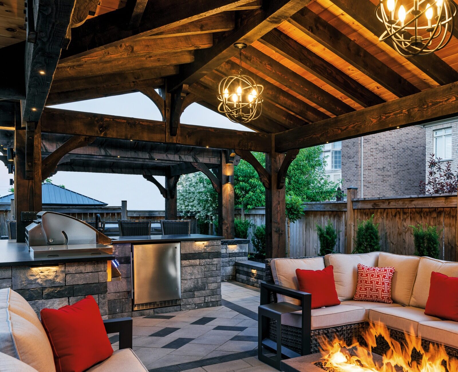 Cozy outdoor patio with wooden pergola, modern grill, and stone fire pit surrounded by beige seating with red cushions and decorative lighting.