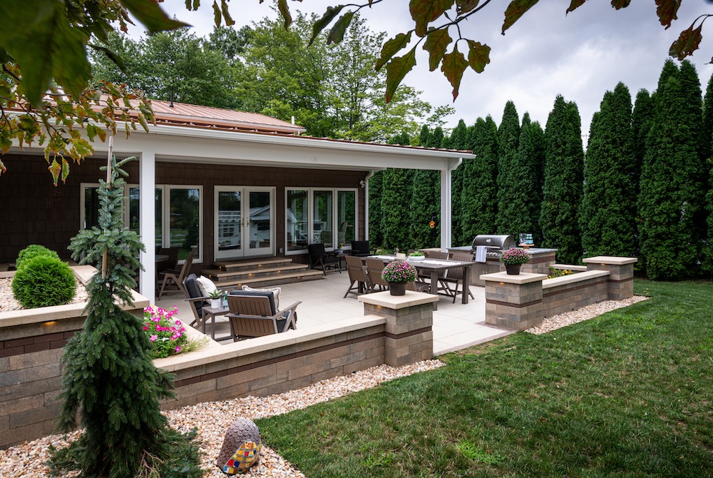 A tranquil backyard patio features seating, flowers, and a barbecue grill, surrounded by lush greenery and trees under a cloudy sky.