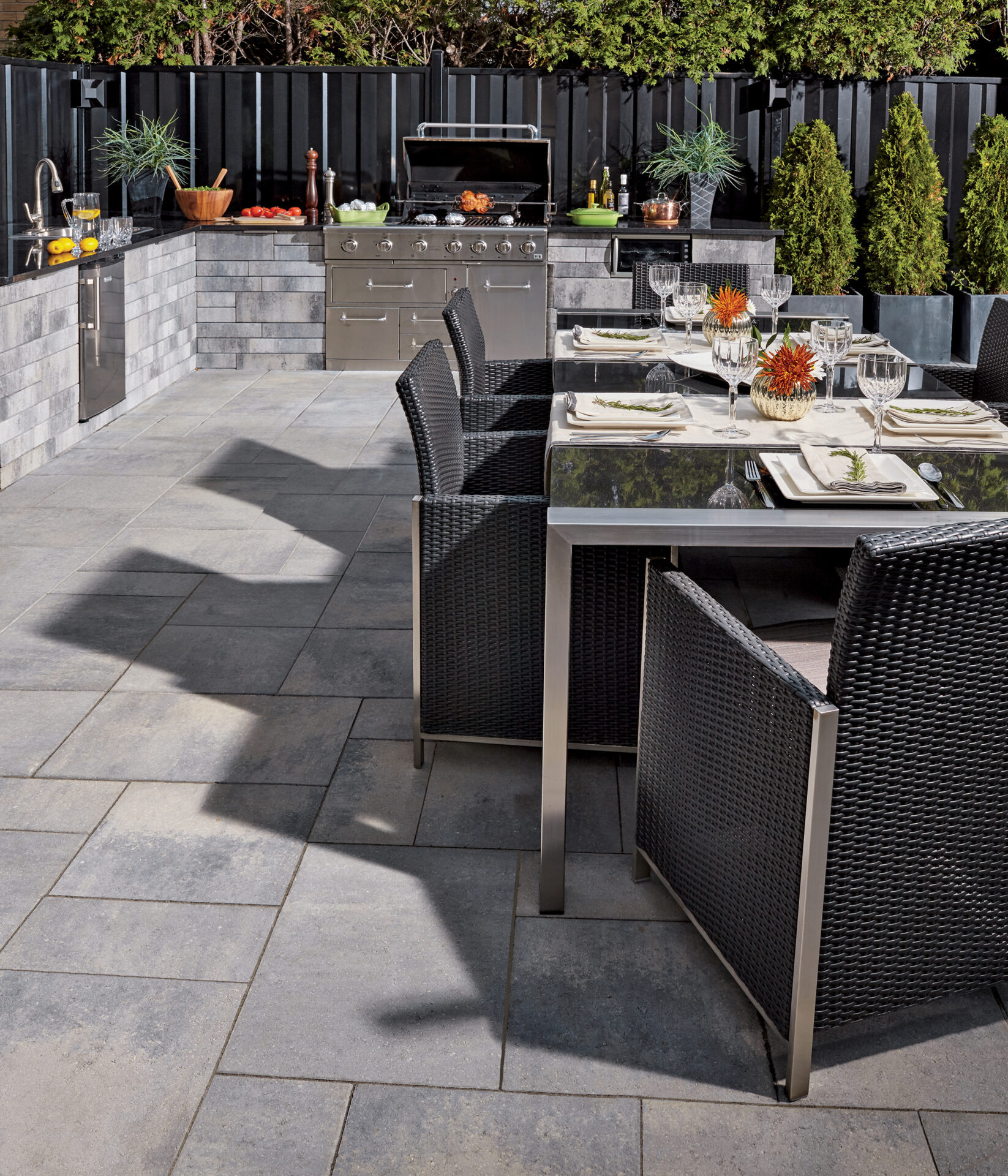 Modern outdoor kitchen with stainless steel grill, sink, and wicker dining set. Elegant table setting, surrounded by greenery and slate tiles.