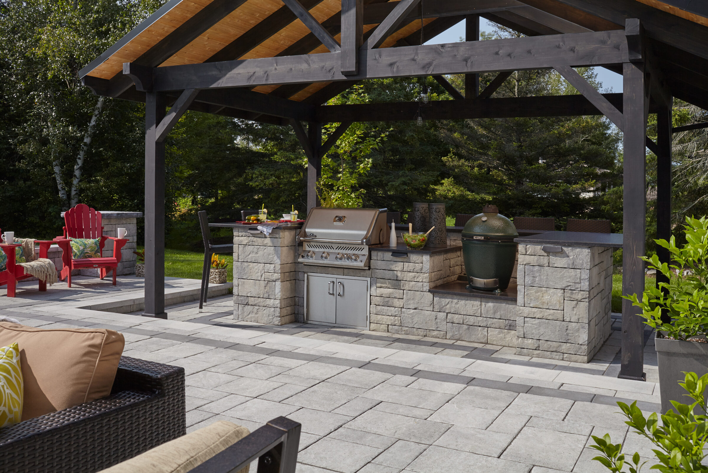 Outdoor kitchen with stone countertop, grill, and seating area under a wooden pergola. Greenery surrounds a relaxing patio space.