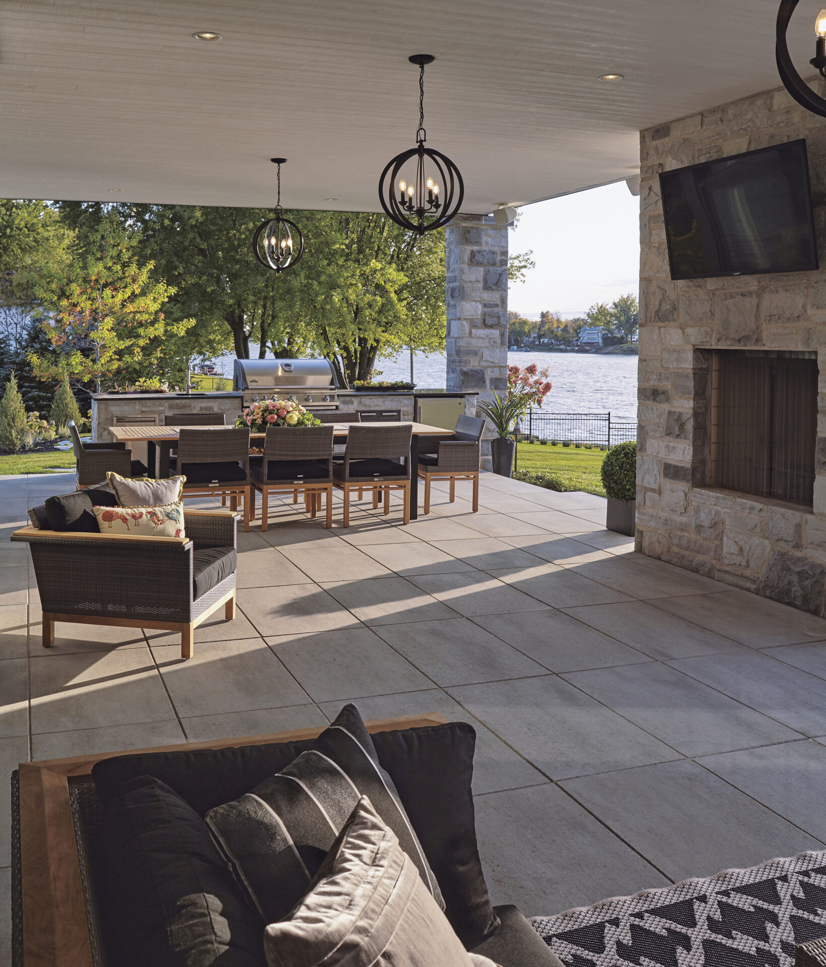 Covered patio with seating, dining table, and grill overlooking a lake. Brick fireplace, modern lighting, surrounded by trees on a sunny day.