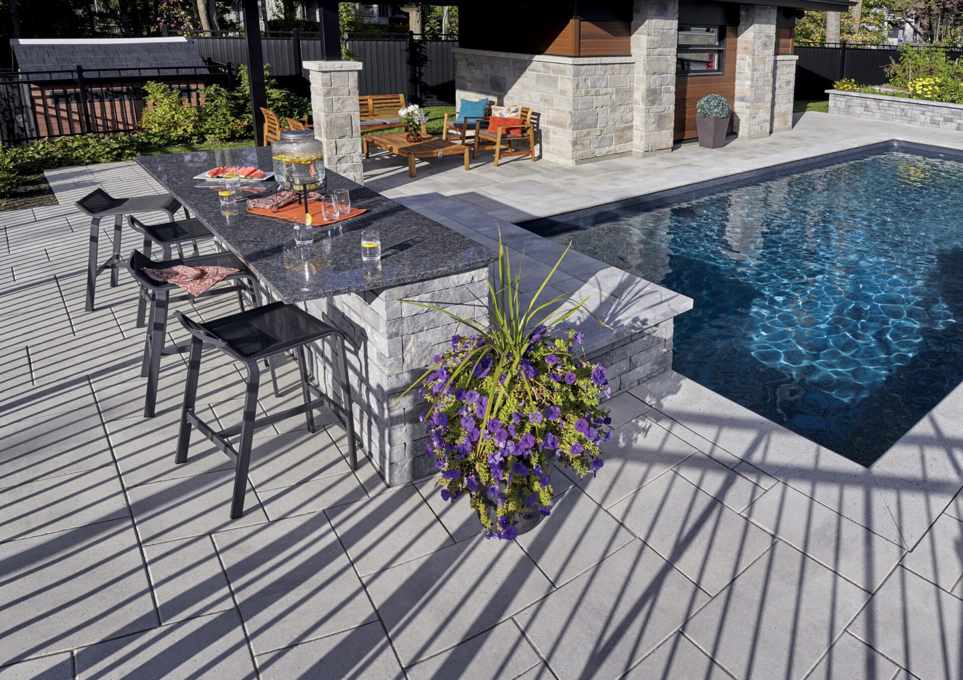 A modern outdoor patio features a pool, stylish bar area with stools, potted purple flowers, and wooden seating under shaded pergola.