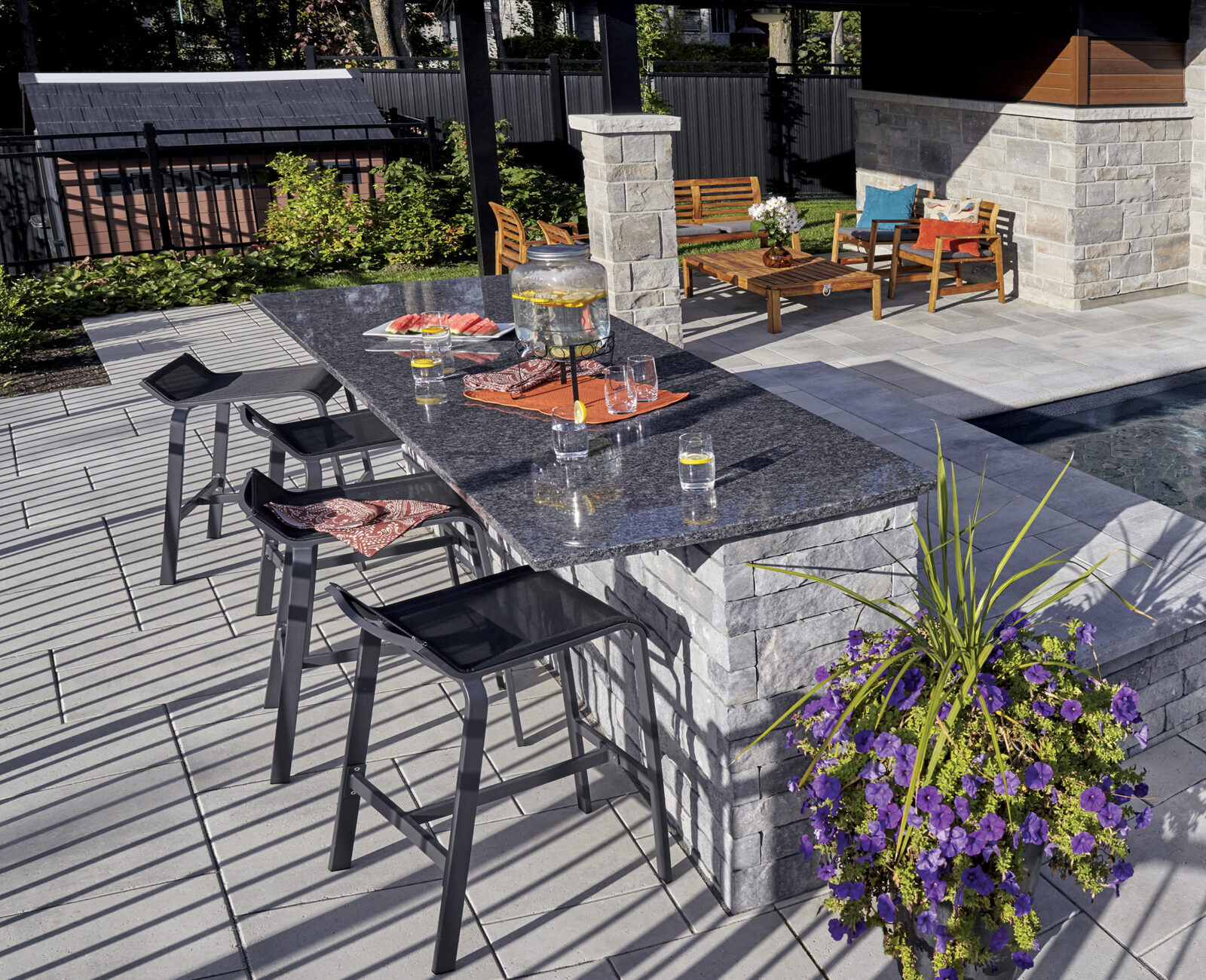 A modern outdoor patio features a pool, stylish bar area with stools, potted purple flowers, and wooden seating under shaded pergola.