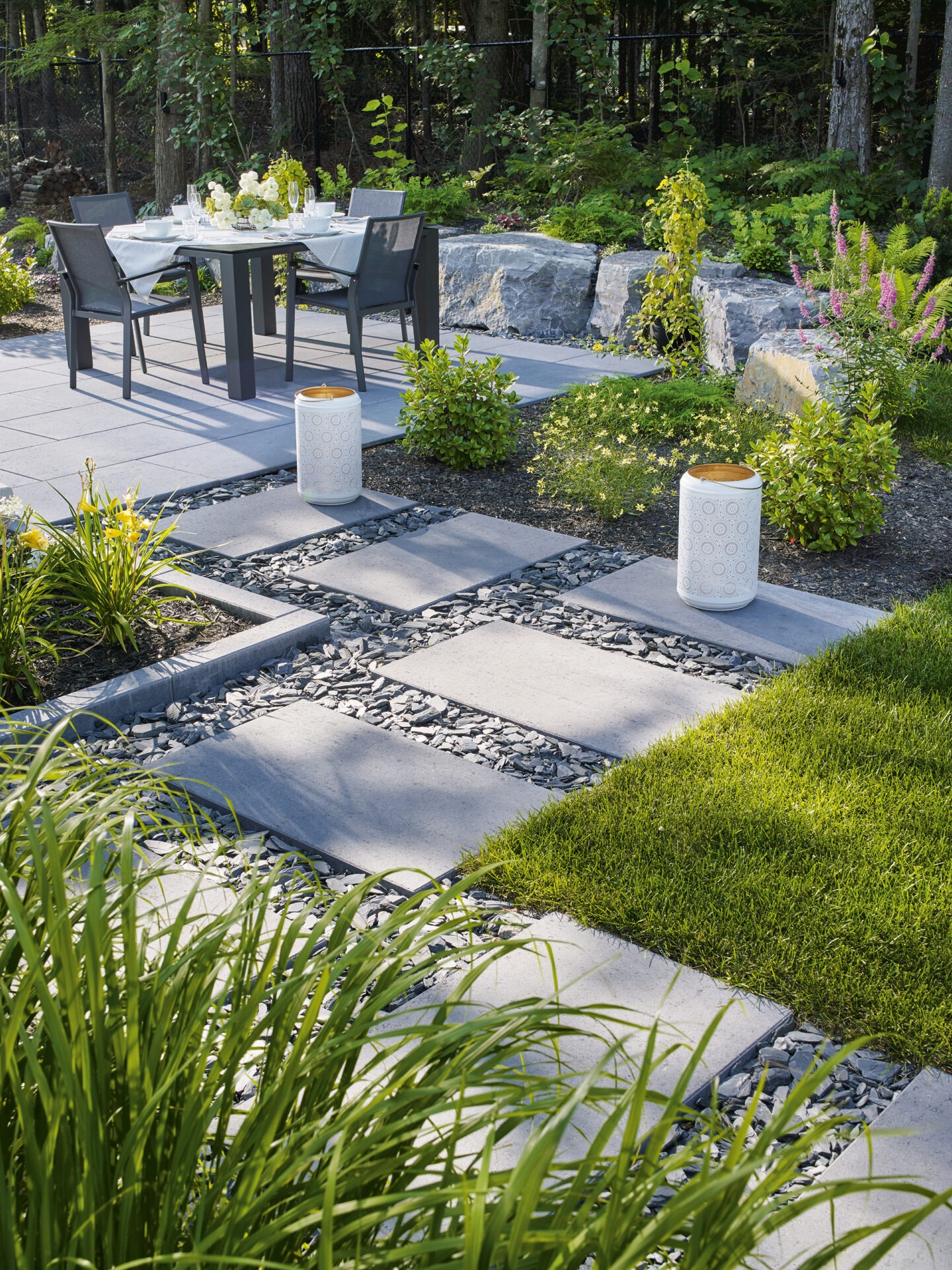 Modern garden patio with dining table set for four. Surrounded by greenery, stone pavers, and decorative lanterns. Serene outdoor atmosphere in lush setting.