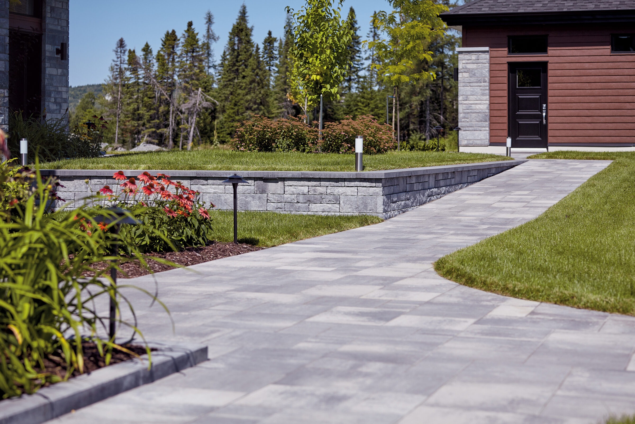 A well-maintained garden pathway with stone tiles, colorful flowers, green lawn, and modern lighting, set against a backdrop of trees and building.