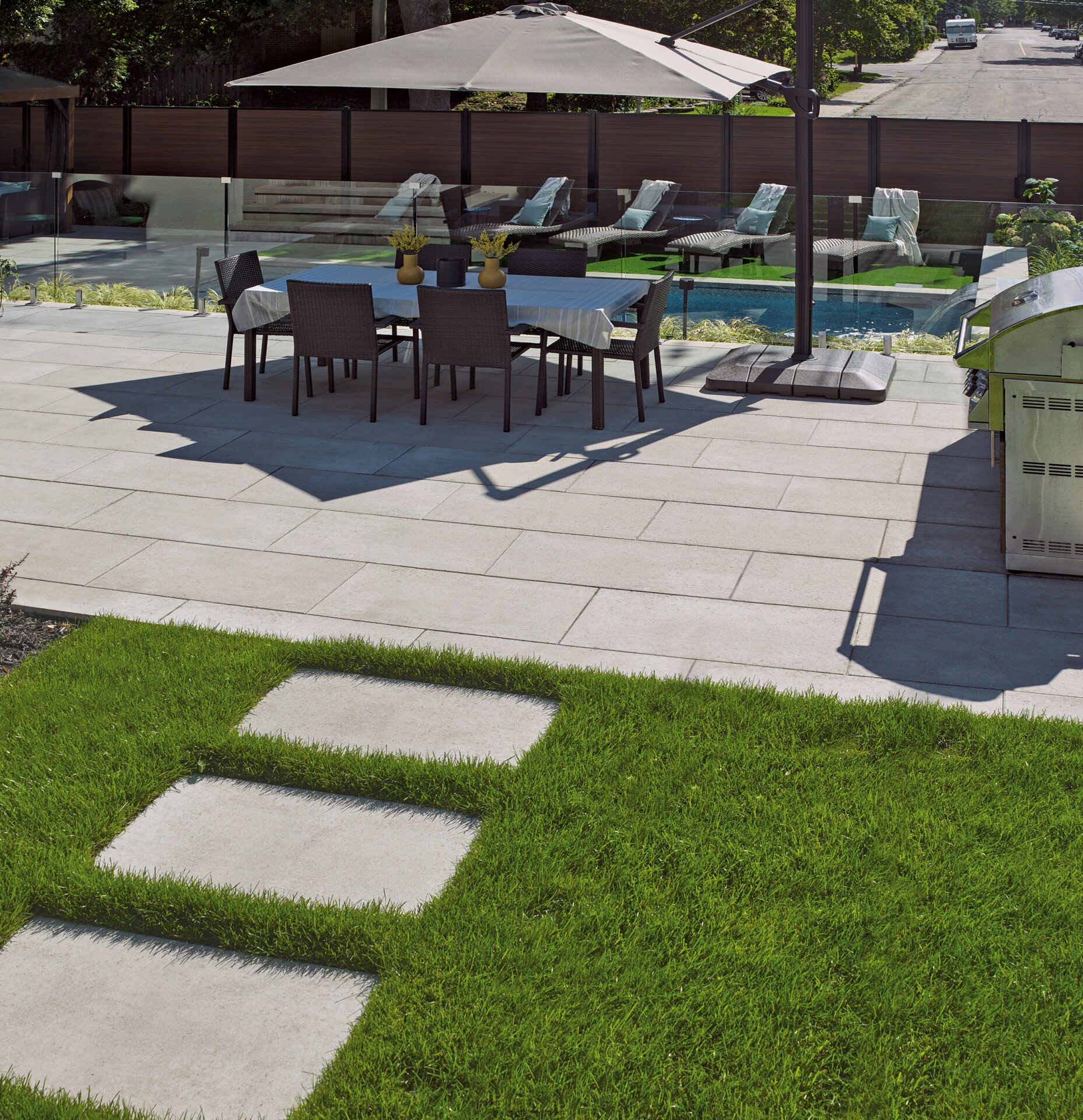 Patio with dining set under umbrella, adjacent to pool with lounge chairs. Green lawn with stepping stones, and street view in background.