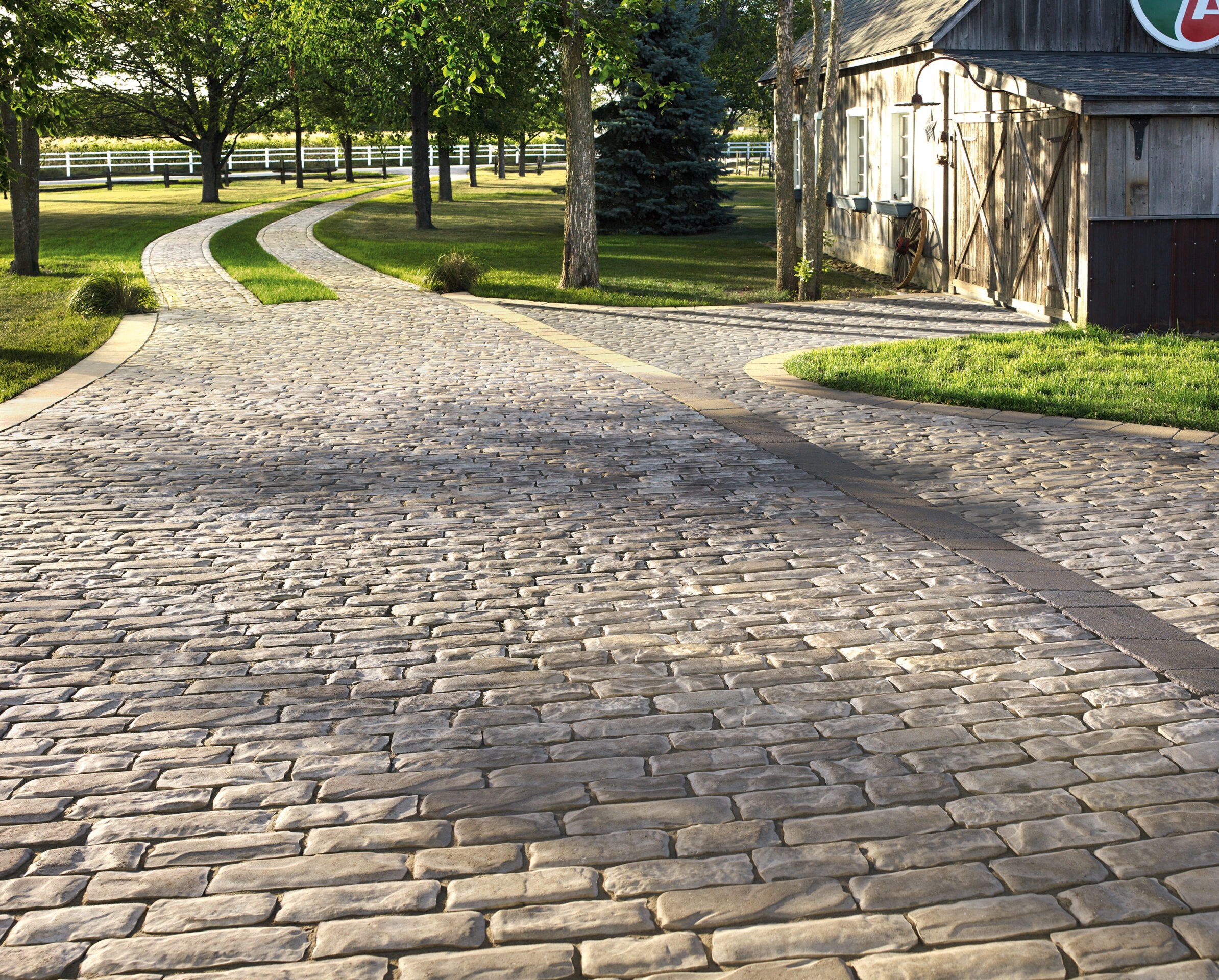 Cobblestone path curves through a tree-lined yard, beside a rustic building with wooden siding and white trim, under a clear blue sky.