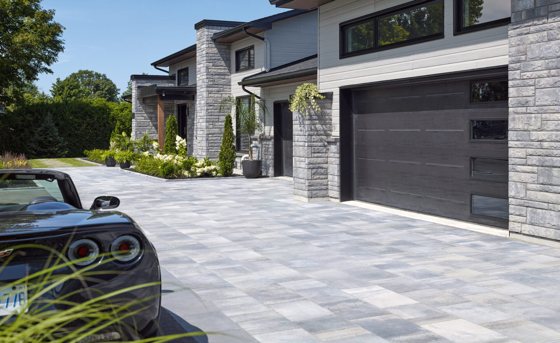Modern stone house with large driveway, black garage doors, and neatly landscaped garden. Convertible car parked in front. Clear sky and lush greenery.