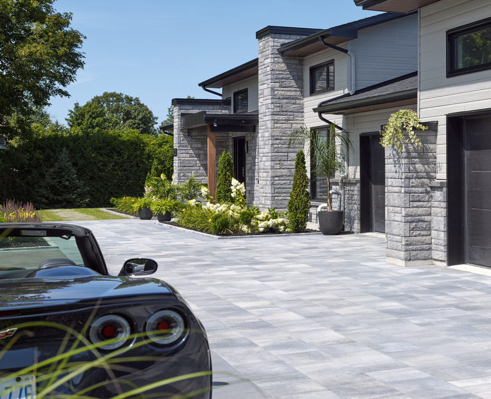 Modern stone house with large driveway, black garage doors, and neatly landscaped garden. Convertible car parked in front. Clear sky and lush greenery.