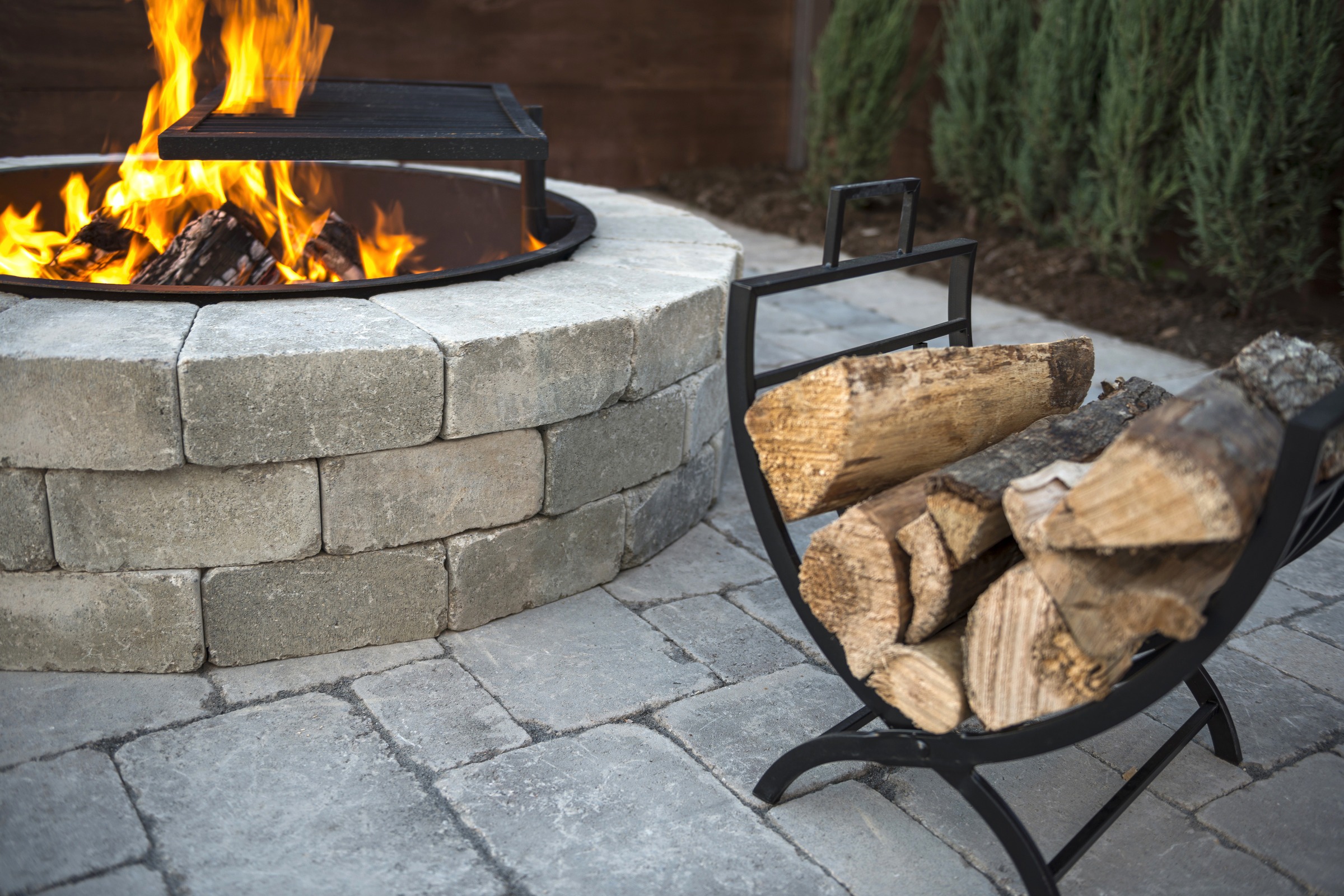 Stone fire pit with burning logs, metal grill, and wood holder on a paved patio, surrounded by greenery, creating a cozy atmosphere.