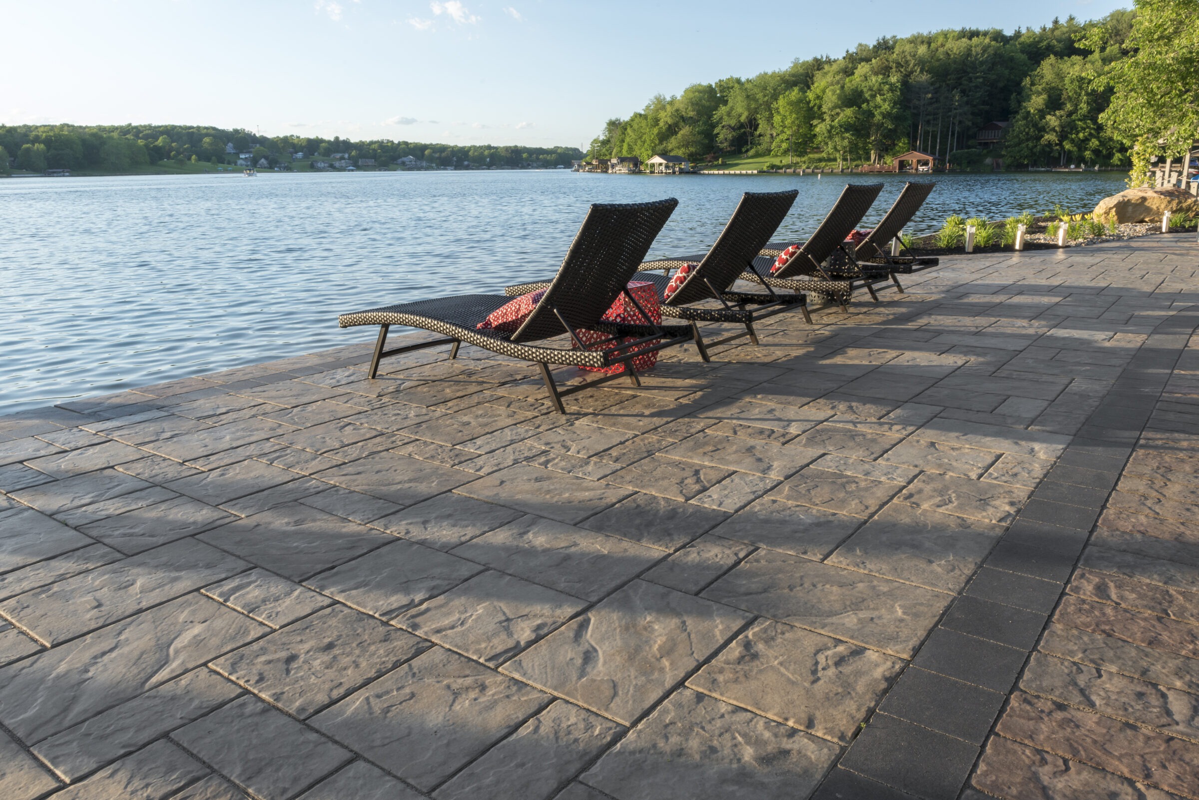 Four empty lounge chairs overlook a serene lake with lush green trees in the background, set on a stone patio by the water.