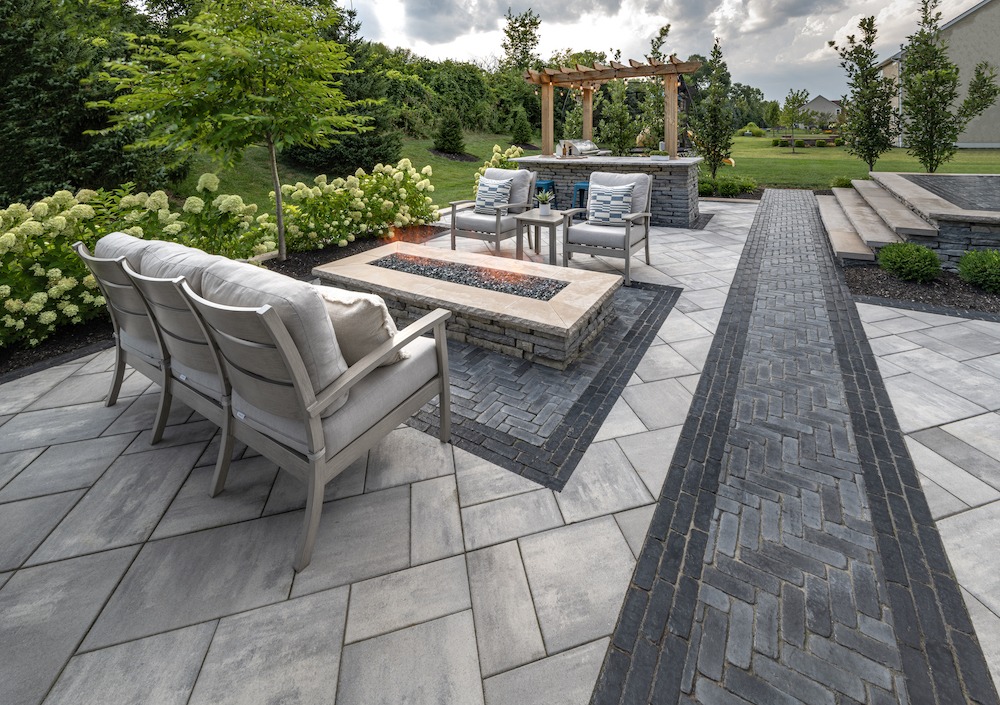 Modern patio with cushioned seating, fire pit, and pergola surrounded by lush greenery. Stone pathway adds elegance to serene garden setting.