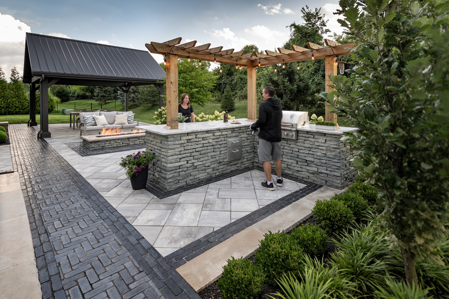 Two people converse in a stylish outdoor kitchen with a pergola, fire pit, and seating area surrounded by greenery and a black-roofed gazebo.