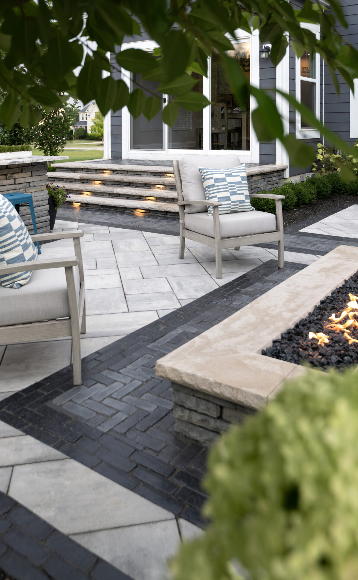 Modern patio with cushioned chairs, stone fire pit, and patterned paving. Foliage partially frames view. No landmark or historical building visible.