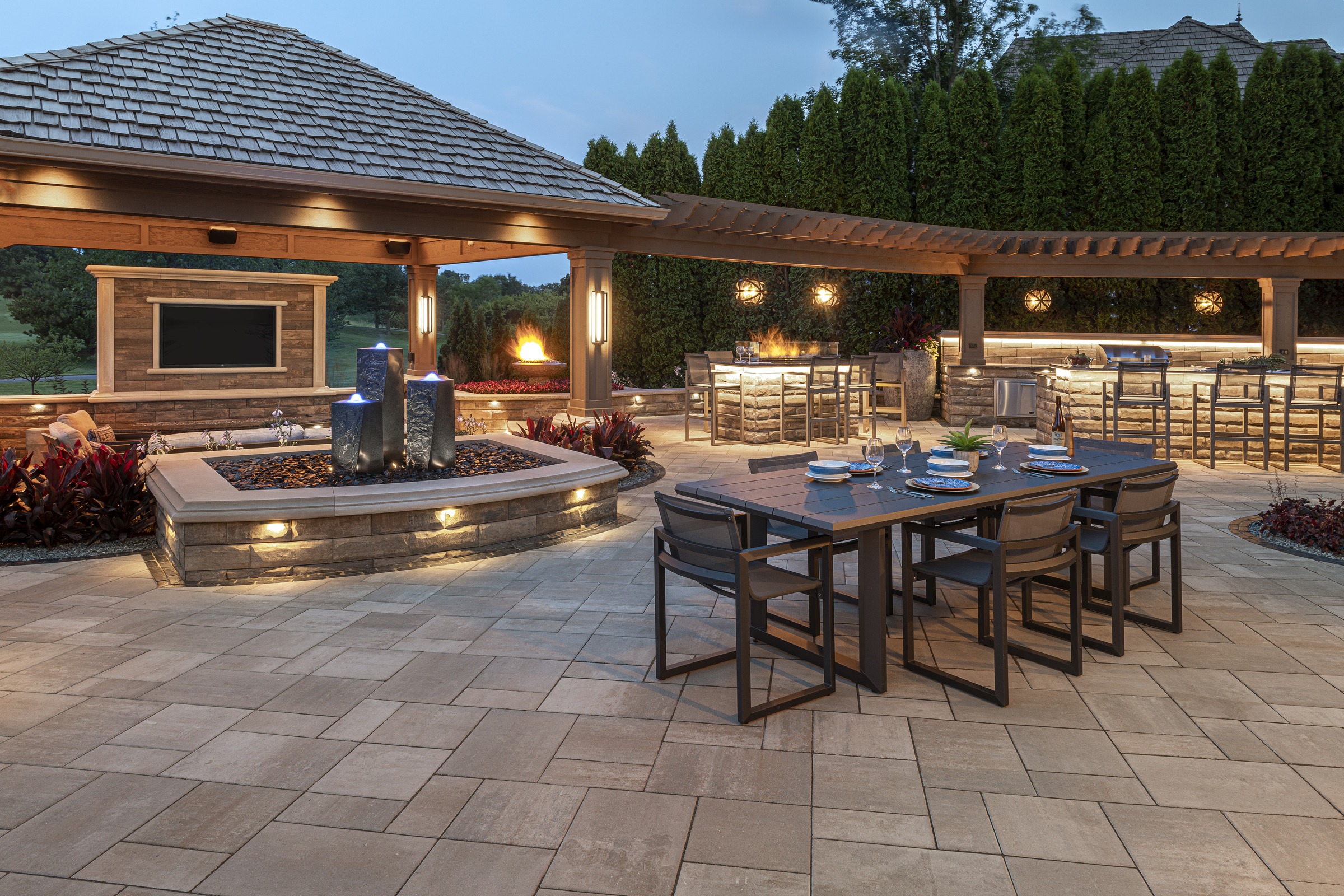 Modern outdoor patio with elegant dining area, water feature, and lush greenery. Soft lighting creates a warm, inviting ambiance in the evening.