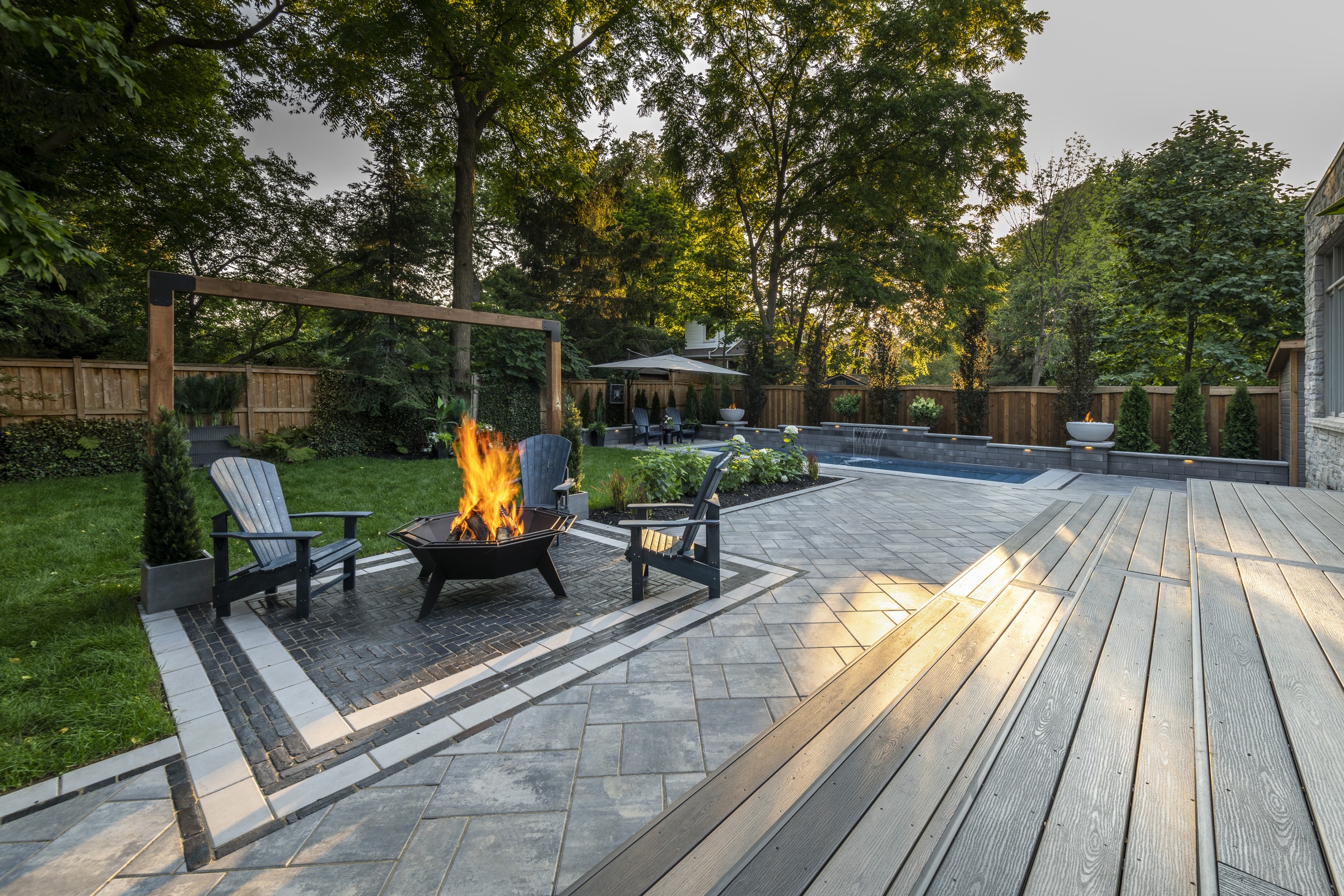 A backyard with a modern patio, fire pit, and Adirondack chairs surrounded by lush greenery and trees, at sunset.