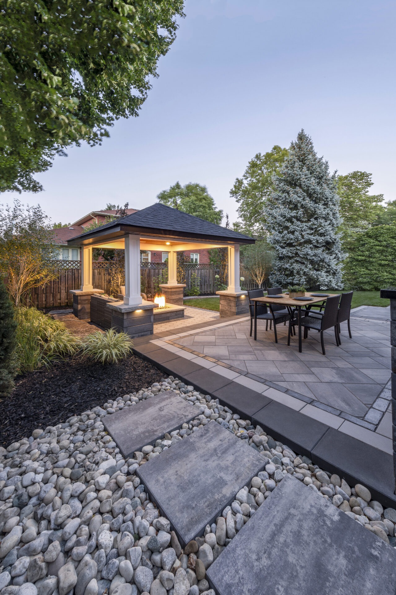 A landscaped backyard features a gazebo with a fire pit, surrounded by trees and a dining area on a stone patio.