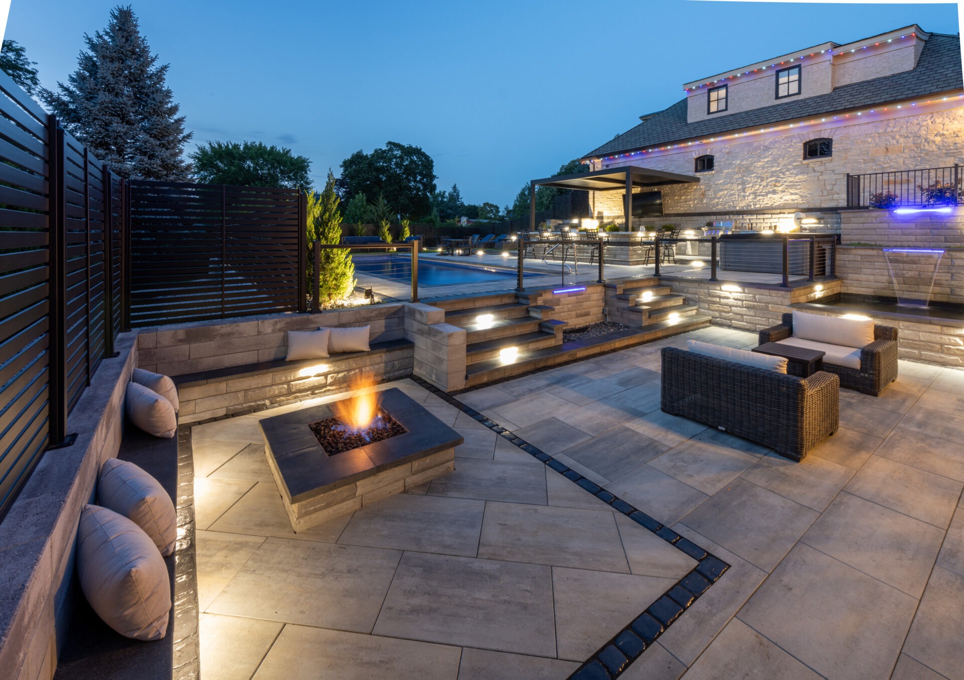 Modern outdoor patio with fire pit, cushioned seating, and illuminated pool area. Elegant stone design and ambient lighting enhance the tranquil evening setting.