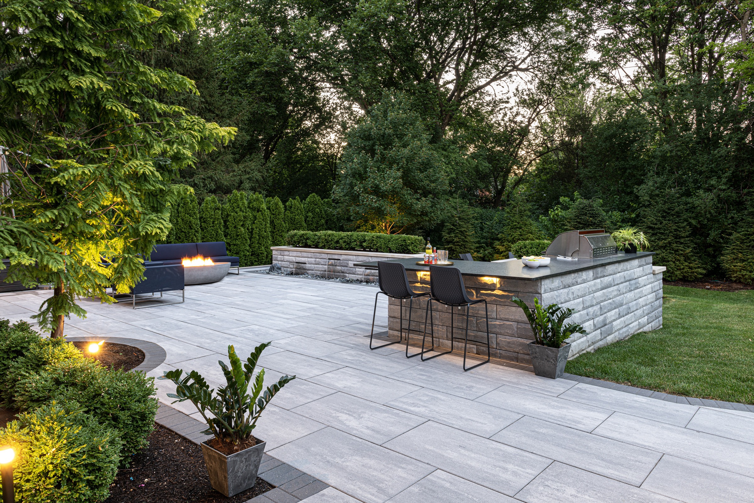 Spacious outdoor patio with modern seating, fire pit, and a stone bar equipped with a grill, surrounded by lush greenery and subtle lighting.
