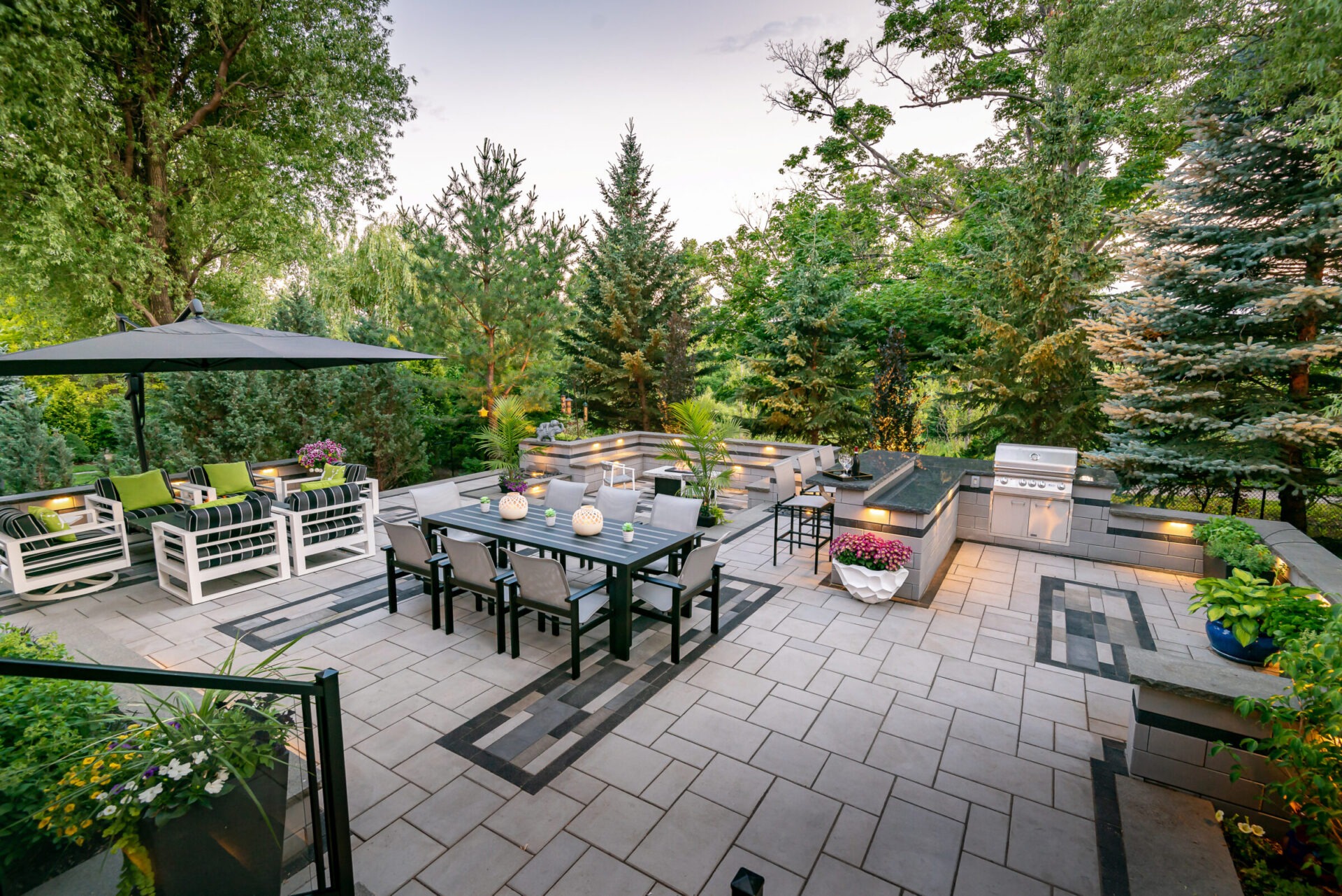 Modern patio with outdoor dining set, large umbrella, and grill. Surrounded by lush greenery, cozy seating, and decorative plants on a tiled surface.