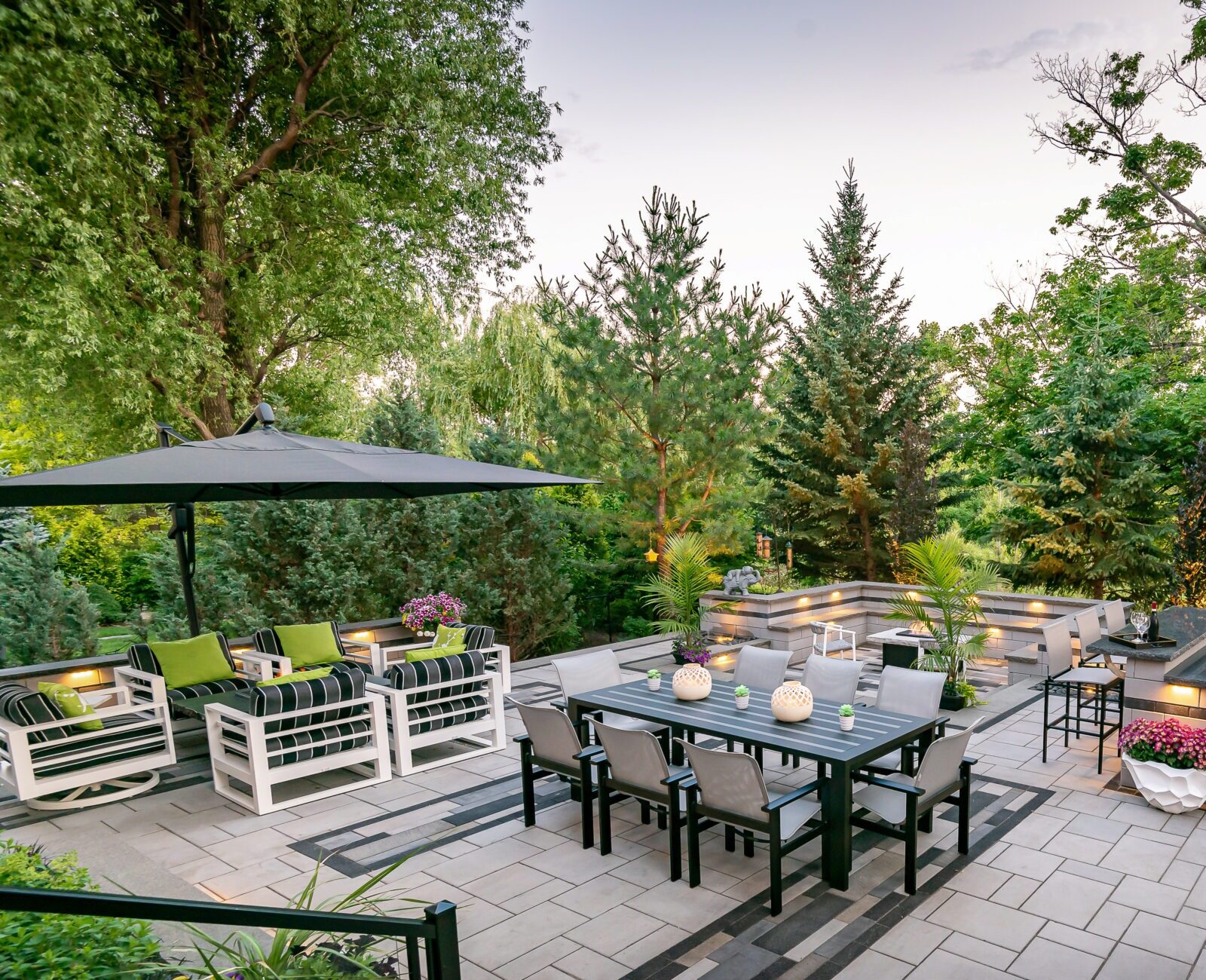 Modern patio with outdoor dining set, large umbrella, and grill. Surrounded by lush greenery, cozy seating, and decorative plants on a tiled surface.