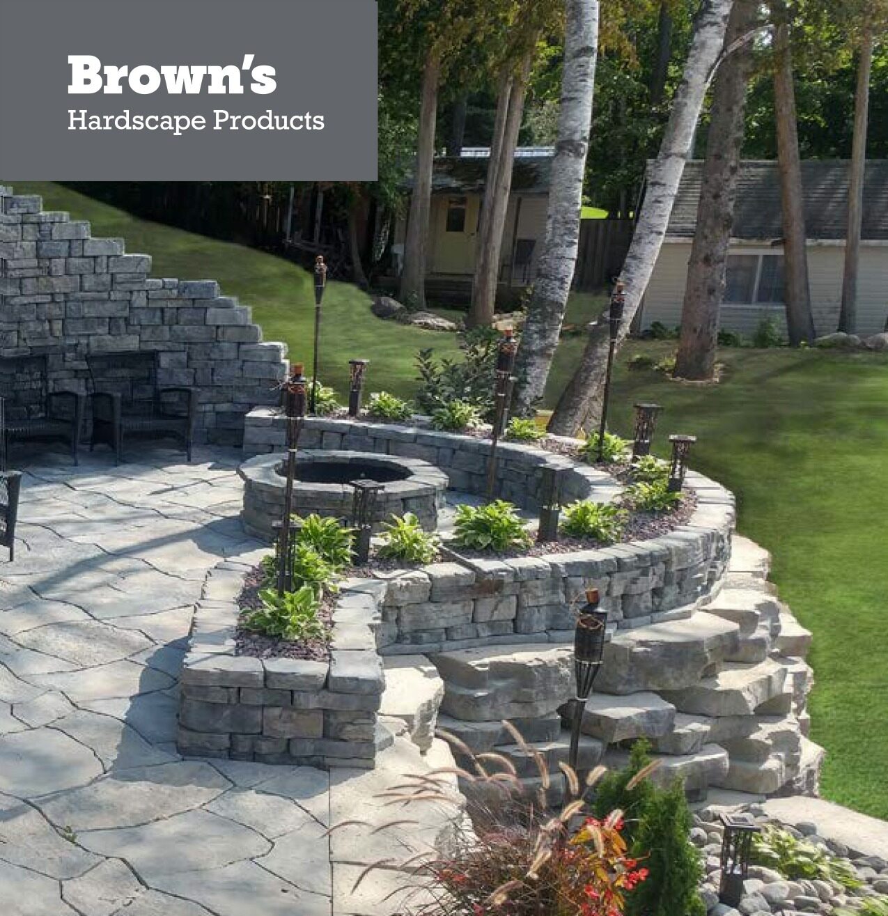 Stone patio with tiered retaining walls, fire pit, and tiki torches surrounded by lush green grass and trees; cozy outdoor seating area.