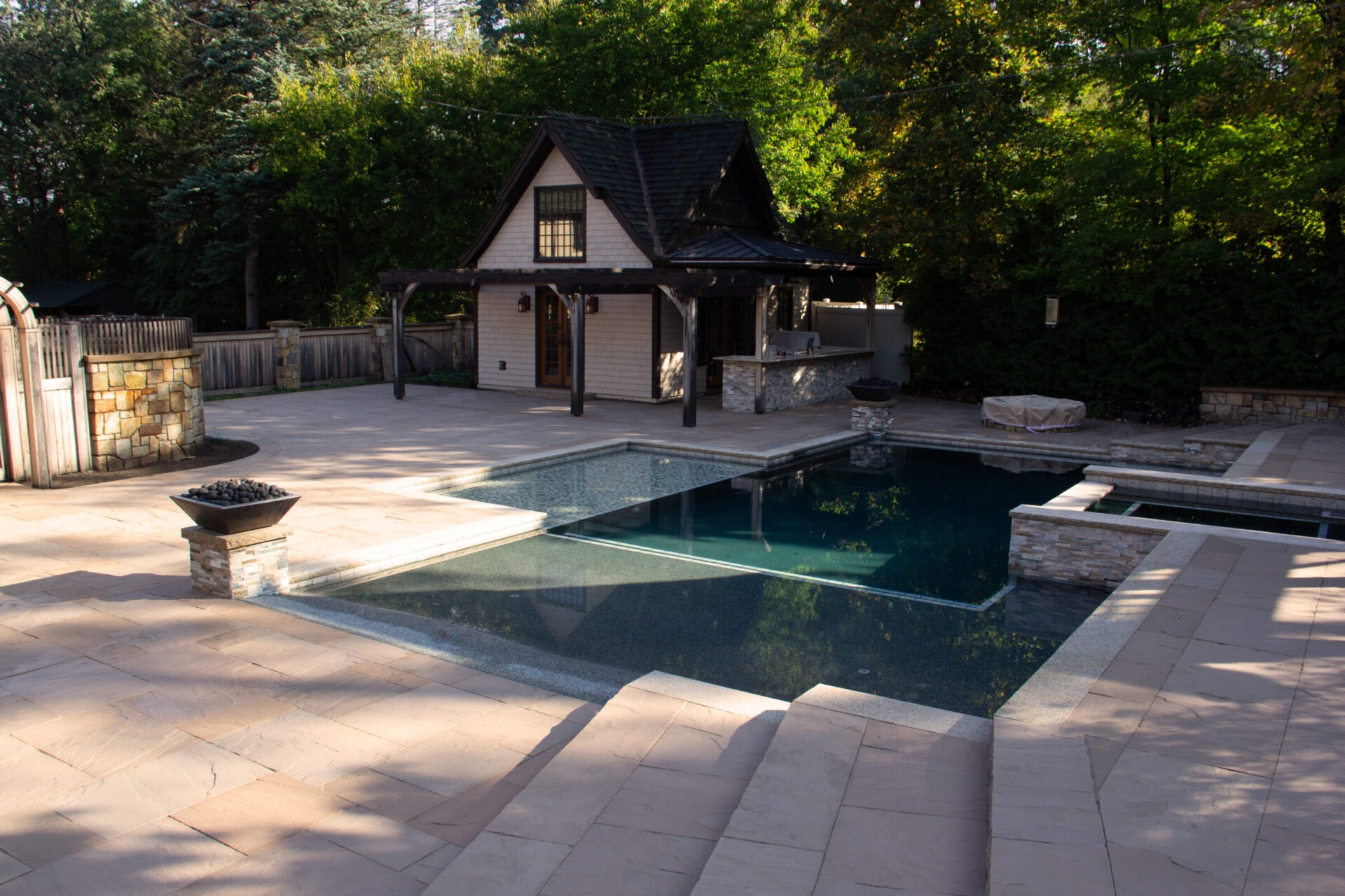 A serene backyard features a modern pool, stone patio, small house, and lush green foliage, creating a peaceful and cozy retreat.