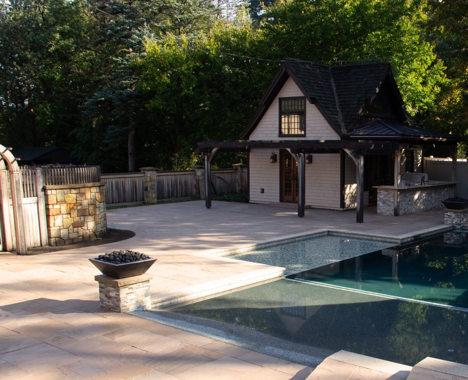 A serene backyard features a modern pool, stone patio, small house, and lush green foliage, creating a peaceful and cozy retreat.