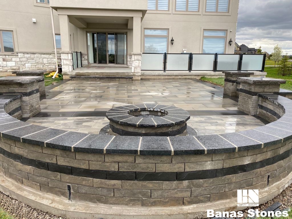 Modern patio with a circular stone design, glass railing, and beige stone house in the background. Overcast skies enhance the landscape's calmness.